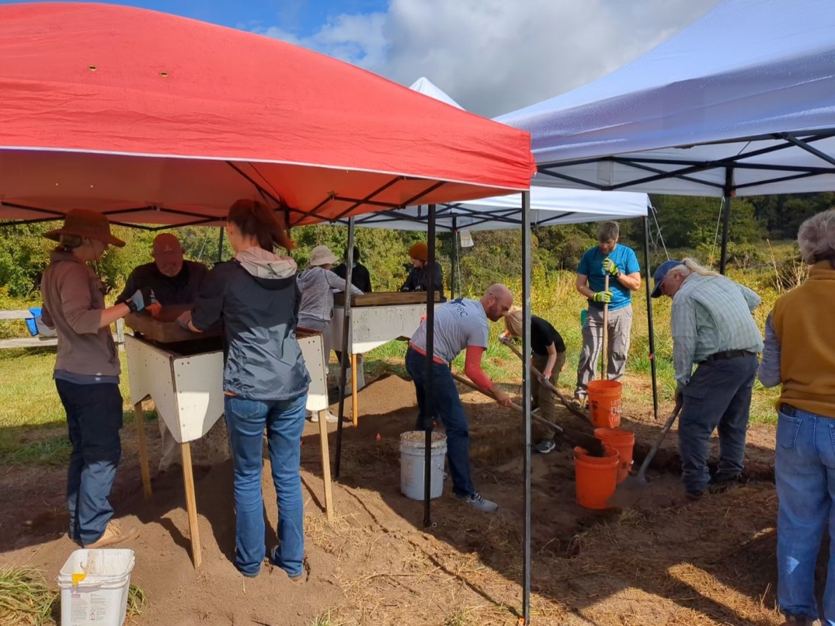 A group of people under pop-up tents against a backdrop of fields and forest shovel dirt into buckets and look through it using raised screens.