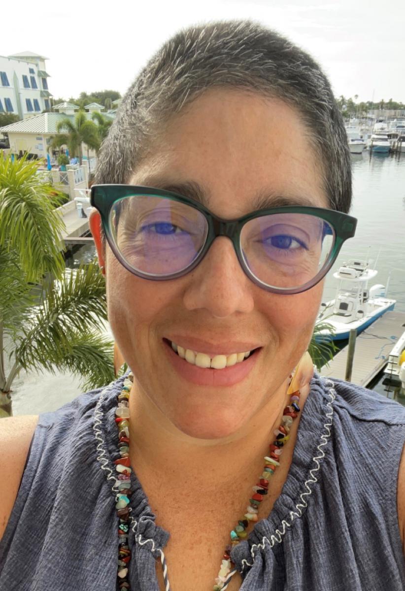 Head and shoulders photo of a woman with light brown skin and short, dark hair, with a marina and palm trees in the background