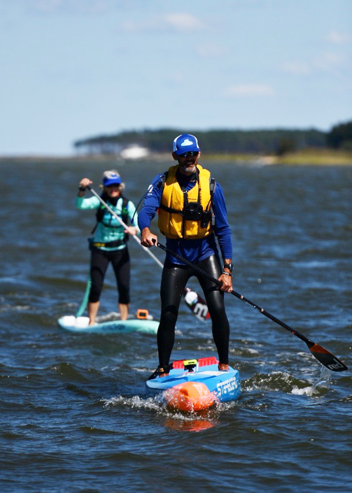 Two people in caps and life preservers paddle on stand-up paddle boards