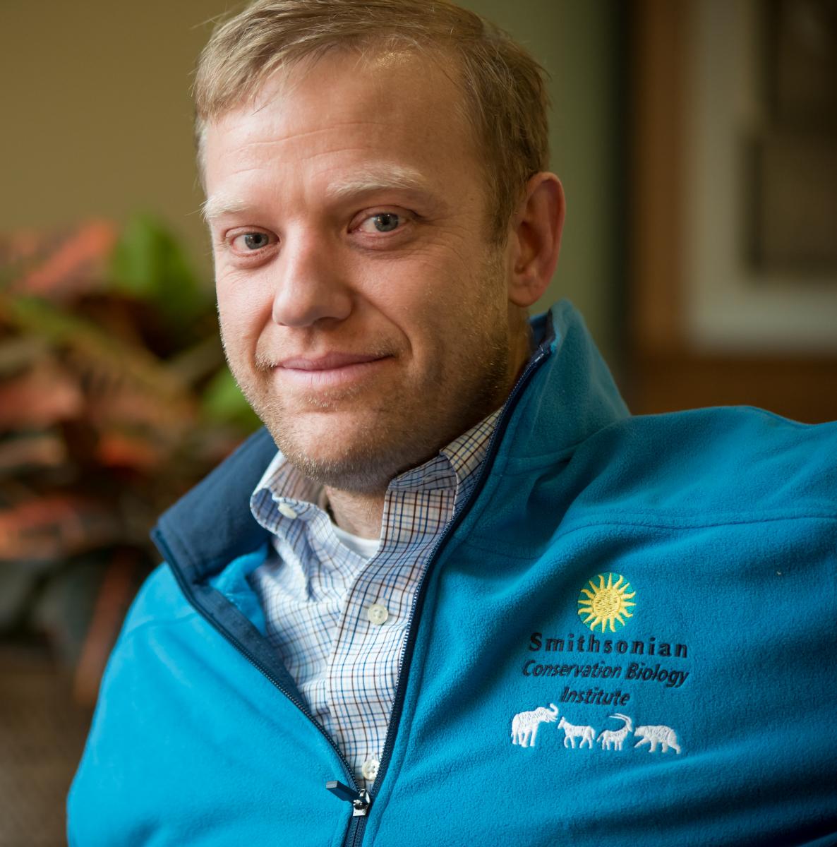 Head and shoulders photo of a man in a blue fleece jacket. Stitched on the right shoulder of the jacket are a blue and yellow sunburst logo, the words Smithsonian Conservation Biology Institute, and the shapes of four mammals in white.