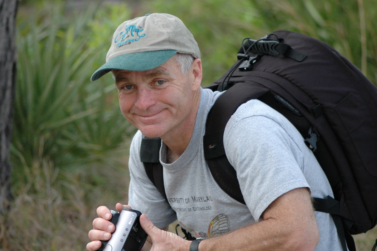 Michael Raupp kneels down in a grassy landscape holding a video camera, wearing a beige cap and black backpack