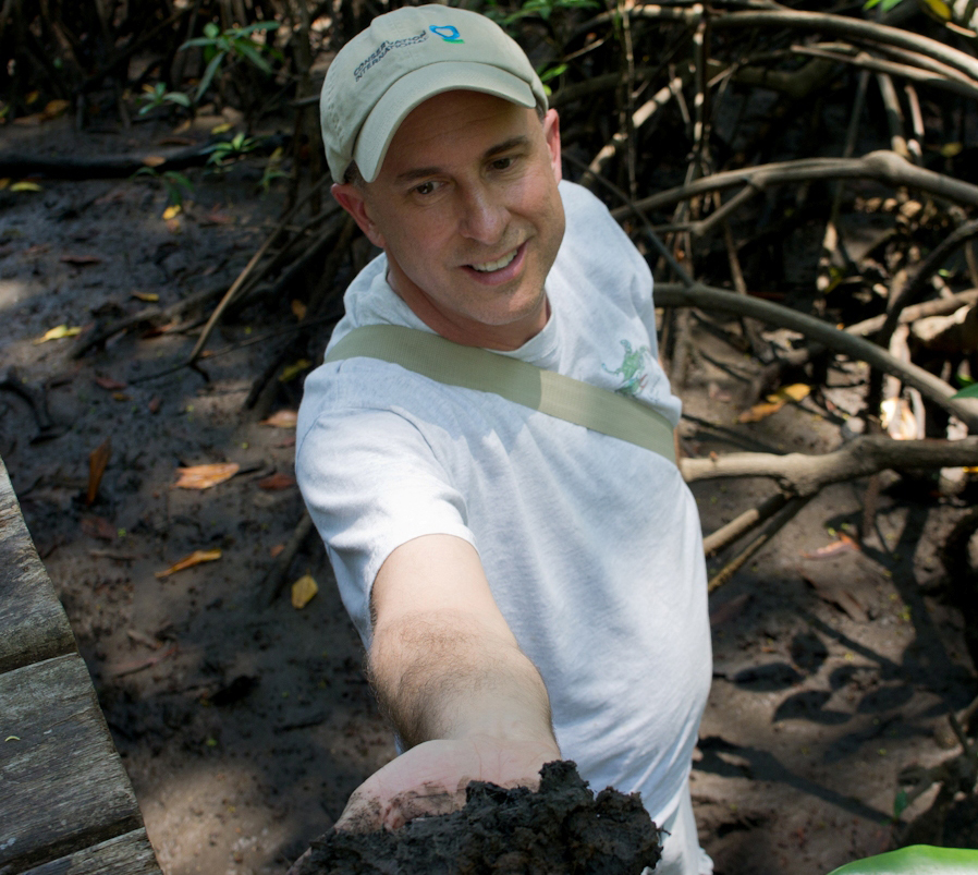 Scientist in whiteT-shirt and beige cap, standing amid tree roots holding a handful of soil up towards the camera