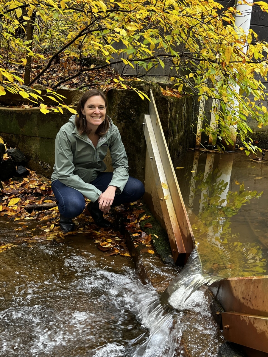 Female scientist wearing a green rain jacket crouches in a stream smiling at the camera, beside a V-shaped weir with water flowing over it.