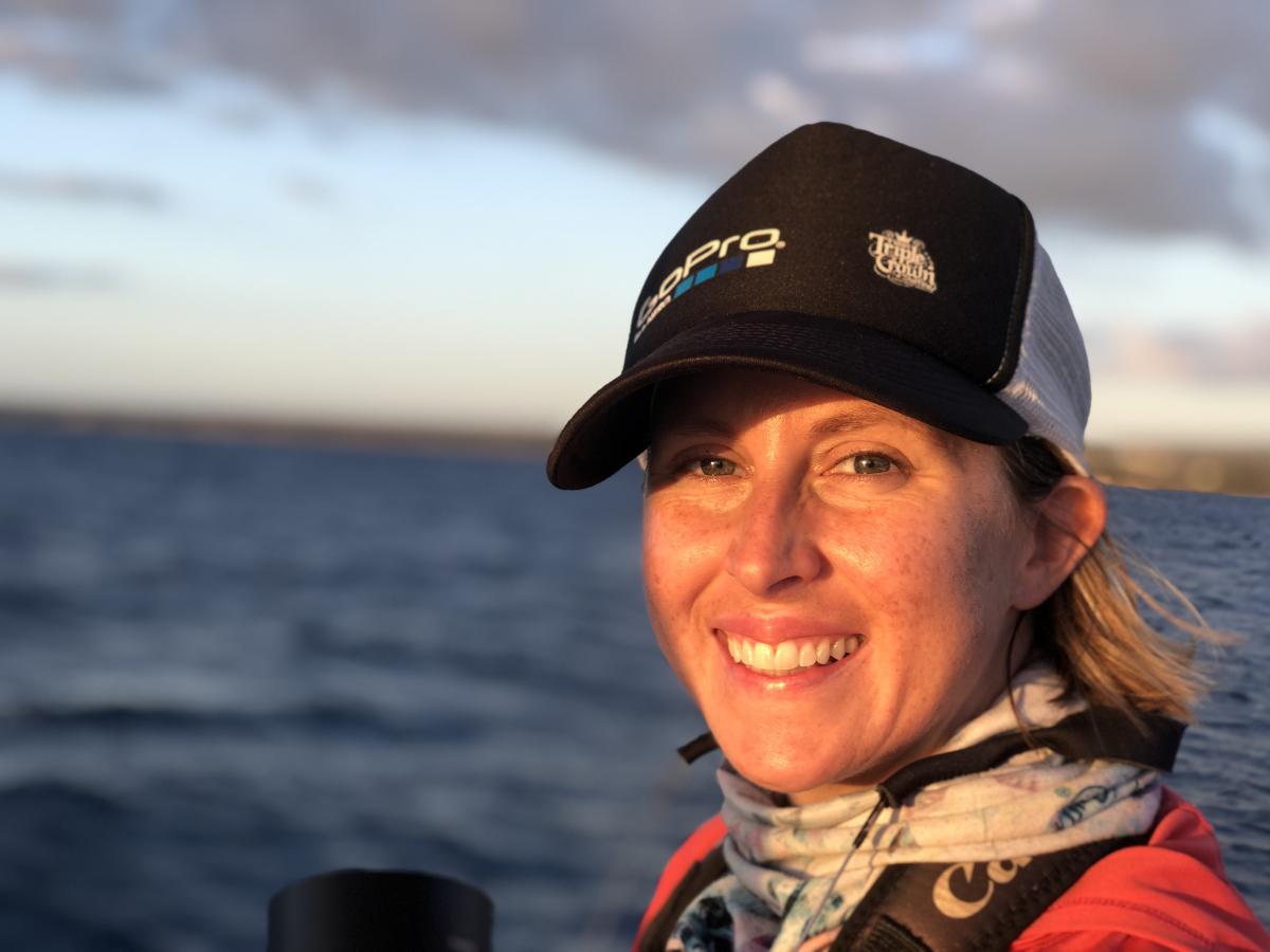 Head and shoulders photo of a young woman wearing a scarf and a black and gray cap, with the ocean behind her