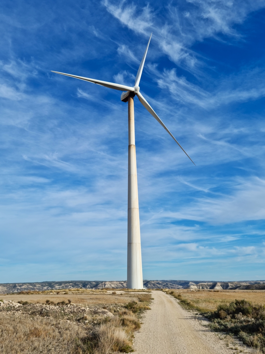 A wind turbine towers above a dirt road, beneath a blue sky streaked with clouds.