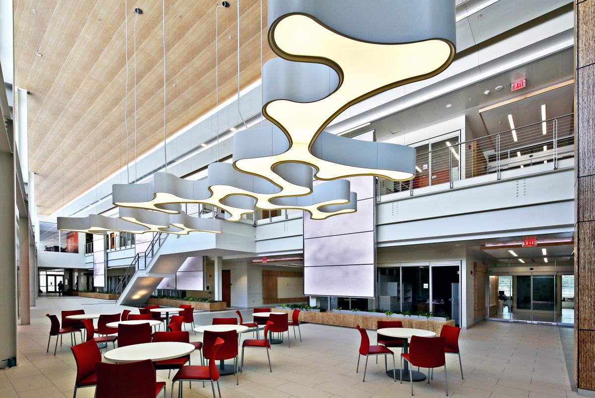 Mathias Atrium table and chairs area, with 7 tables with 4 red chairs each. There is a large asymmetrically shaped light fixture hanging from the ceiling above. The light fixture is silver-colored, and the bottom is yellow-colored from the glow of the light.Behind this seating area is a staircase and several glass doors leading to other offices or outside the building. You can see the second floor overlooking the atrium, enclosed by a silver wired railing.