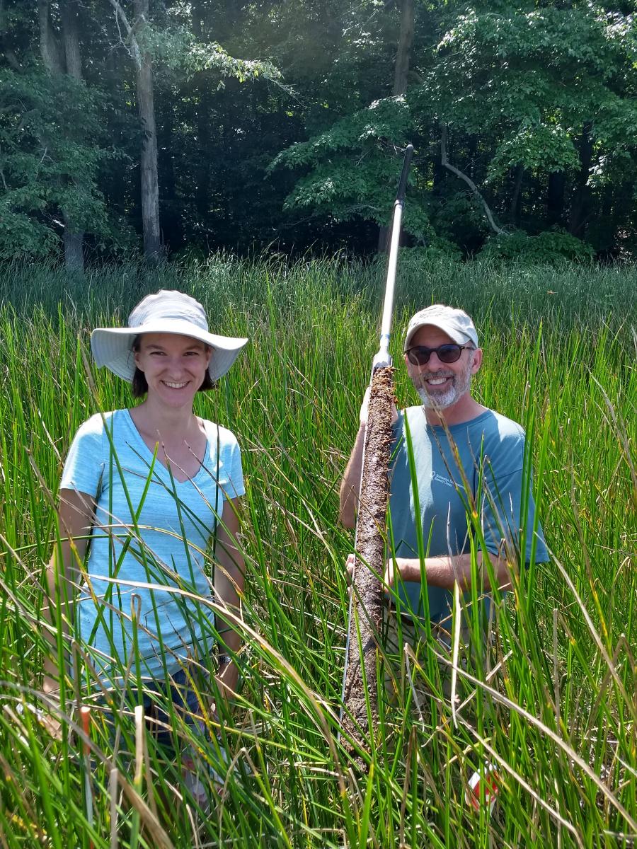 Female and male scientist standing in marsh; man holds soil core