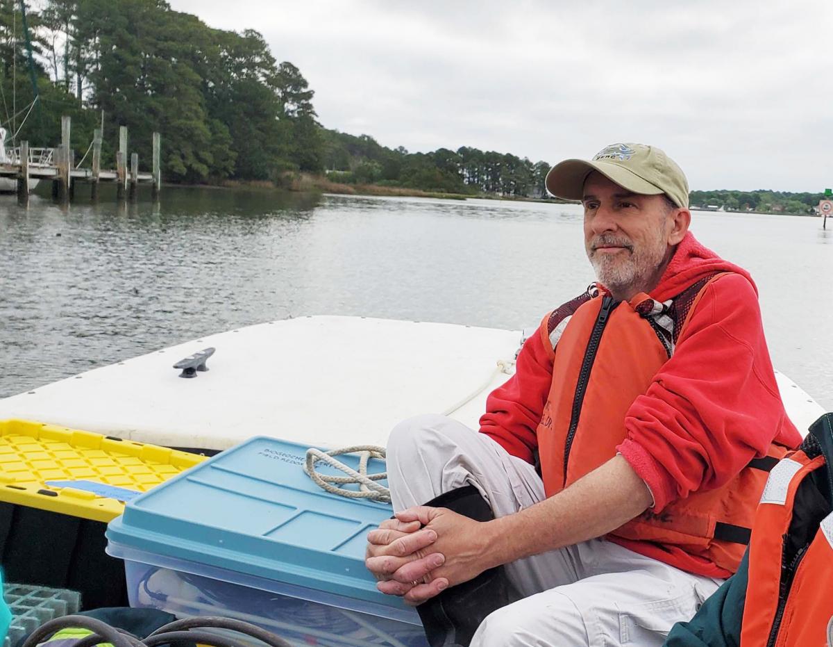 Scientist in a red jacket and beige cap sits on the back of a boat gazing forward, with forested marshy shoreline to his right