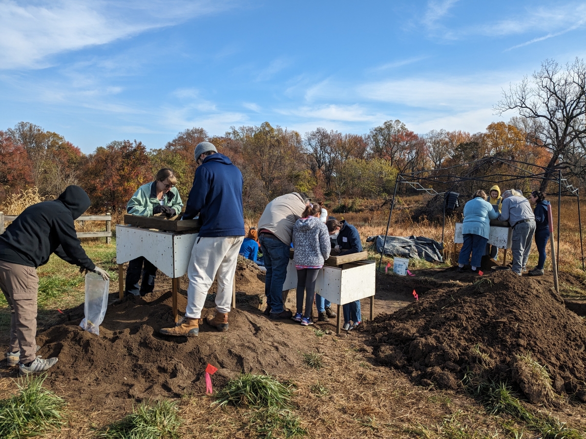 Groups of people gather around three standing screens. Around them are mounds of sifted soil, small field flags. The backdrop is a fall forest.