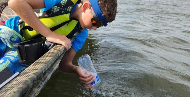 A yov volunteer takes a water sample from a dark blue-green river. from a boat.