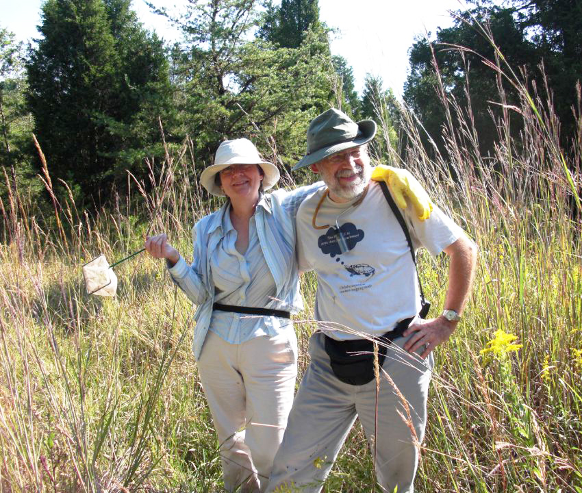 : Charlie and Sue Staines pose for a camera in a grassy field, surrounded by tall reeds. Sue drapes her right arm over Charlie's shoulder, while holding a small bug-catching net in her left hand.