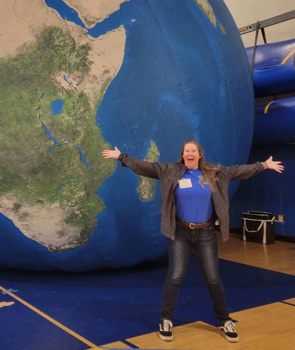 Rachel Terracina wearing a dark gray unbuttoned shirt over a blue shirt, and jeans. Rachel is posing in front of a large globe with her arms spread apart.