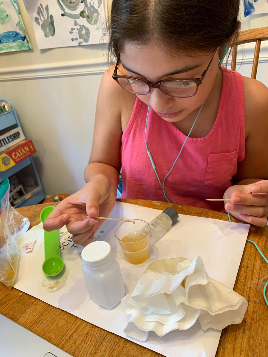 Young girl does a science activity at a table with a plastic cup filled with yellow liquid, a paper coffee filter and and two toothpicks