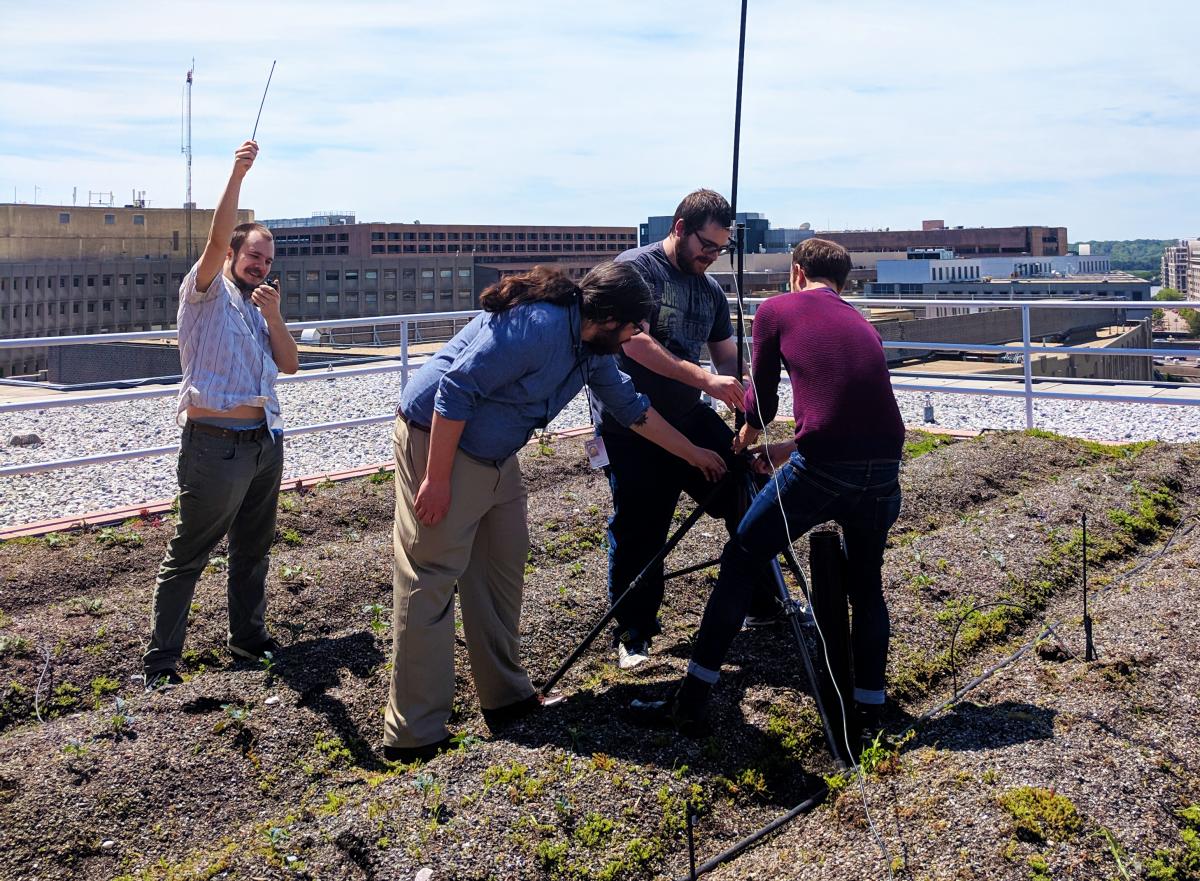 Team of four young adults sets up an antenna on a city rooftop. One of them raises his fist in the air