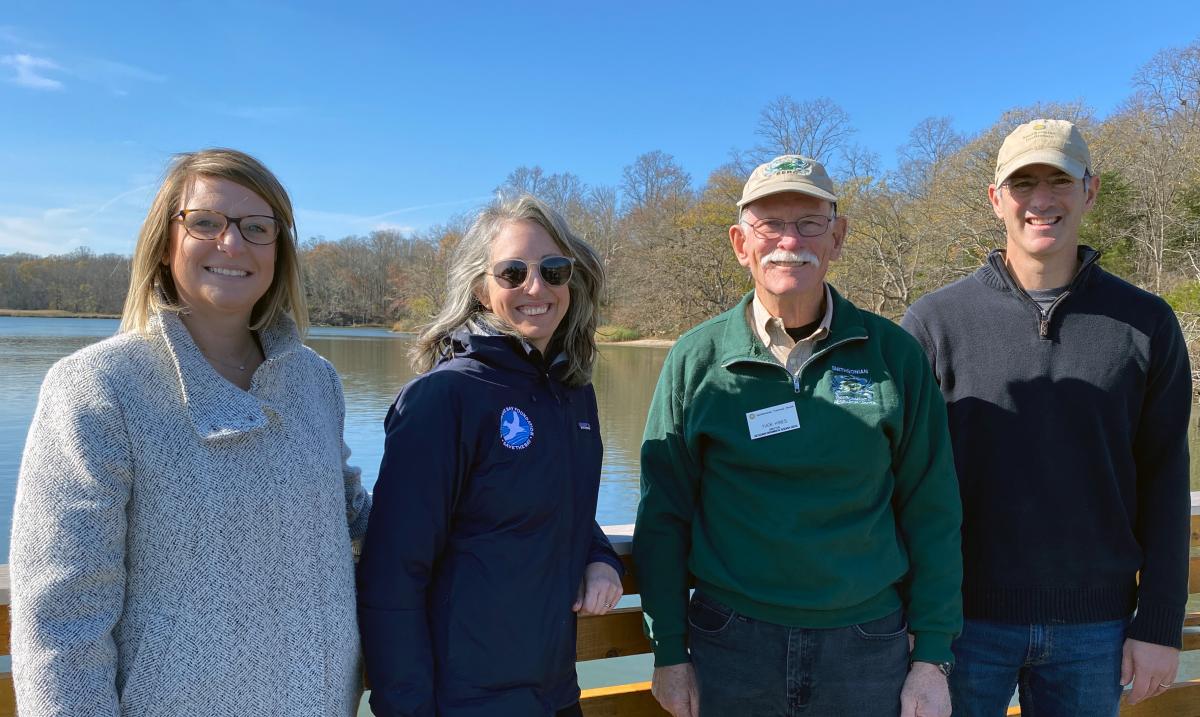 Four people stand on a dock, with a river and a shoreline autumn trees behind them