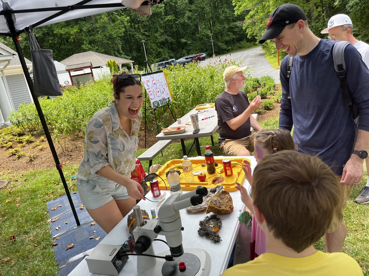 A young woman stands behind an outdoor exhibit table with a microscope, smiling and laughing, while a young man and two children look on.