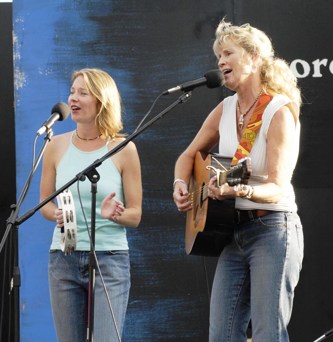 Two blonde women singing on stage with a tambourine and guitar