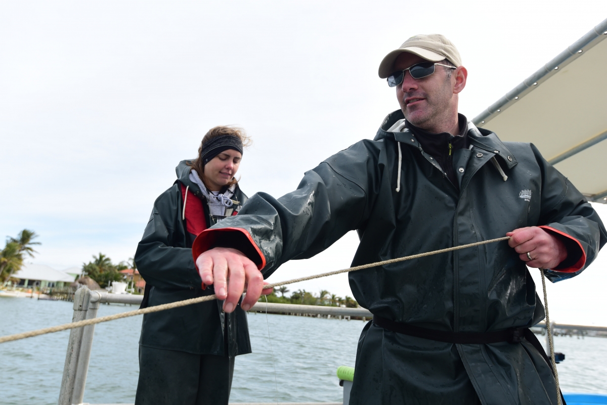 A man in a black jacket with sunglasses and a young woman stand on a boat. The man holds a line of rope in front of him.