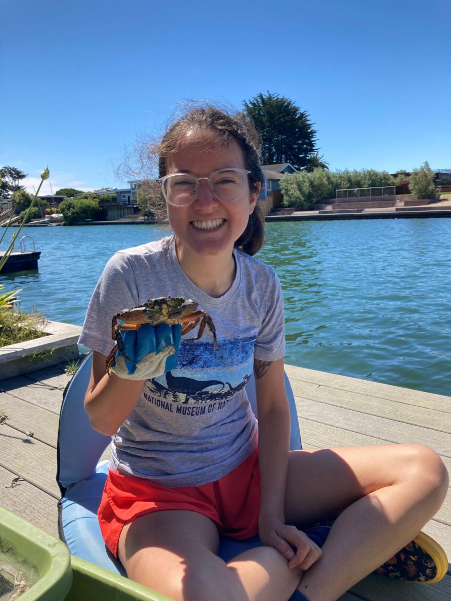 Young woman with light brown skin and glasses kneels on a dock, holding out a green crab