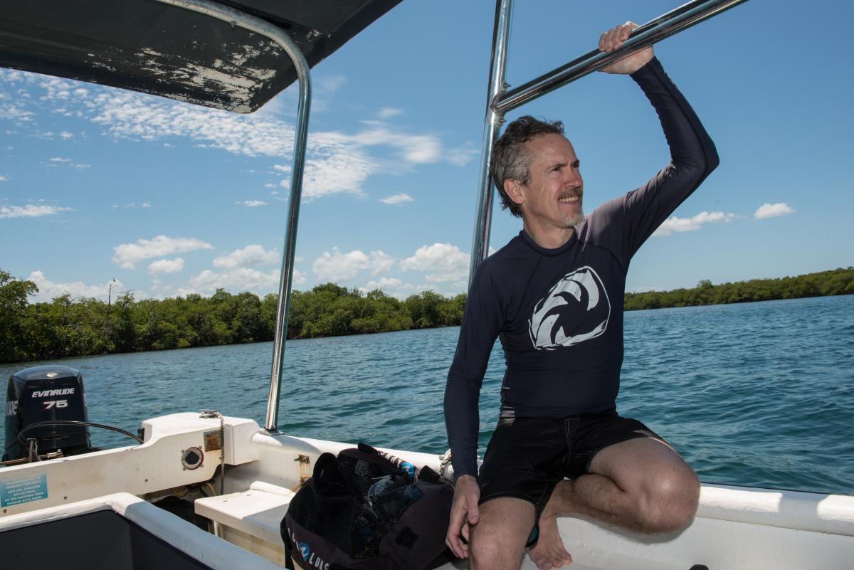 Scientist in long-sleeved swim gear kneels on the side of a moving boat, gazing out at the water