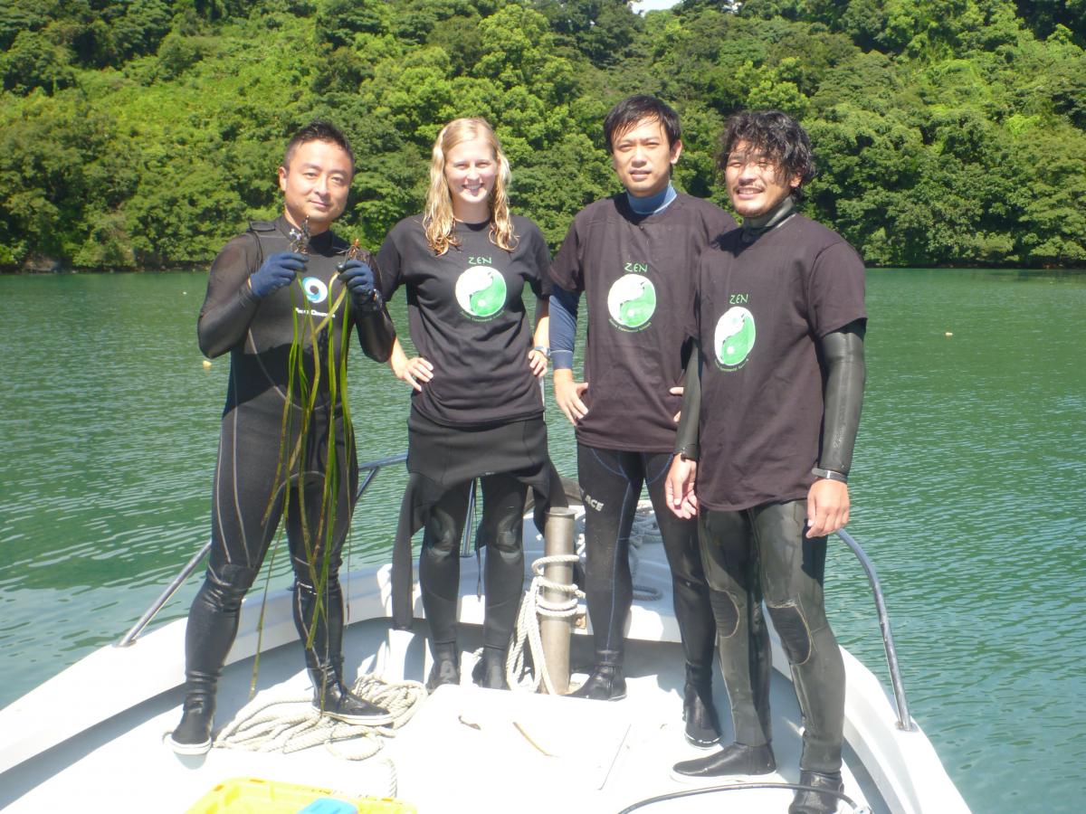 Four people in wet suits and black T-Shirts with the ZEN logo stand on a boat. The one on the left holds up strands of seagrass in both hands