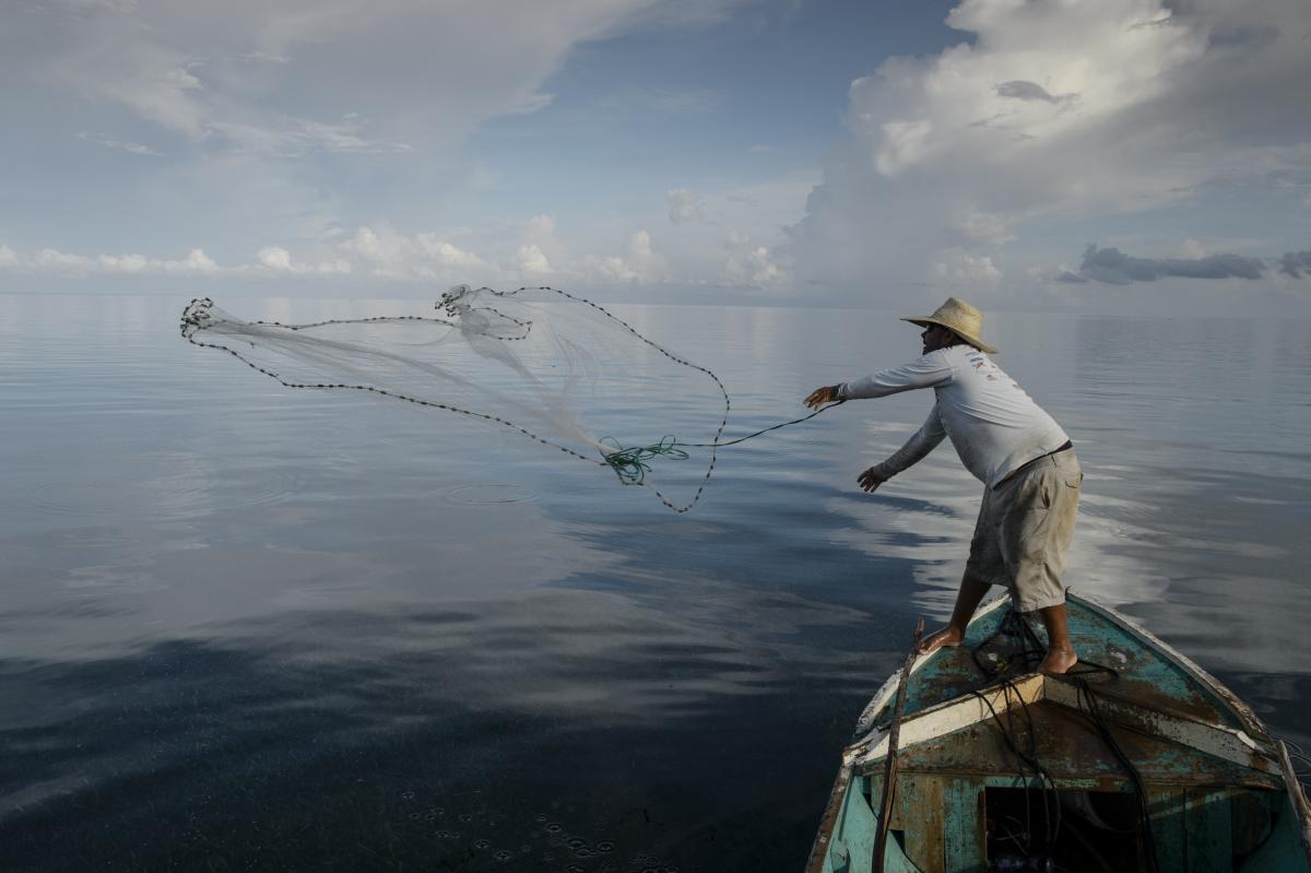 Man on boat wearing straw hat throws a fishing net into the water