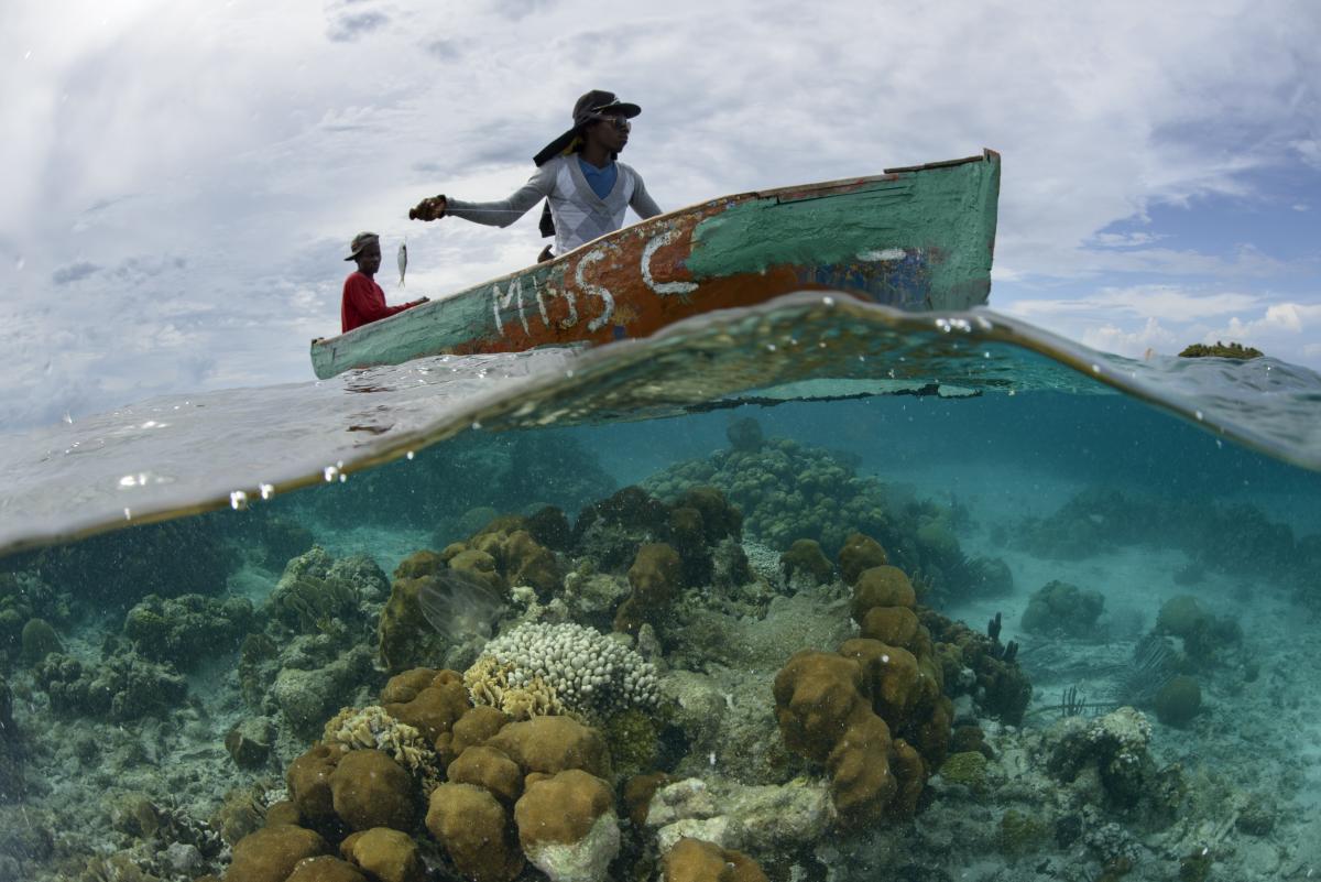 Two dark-skinned people sit in a small green boat, one holding a small fish on a line. The lower half of the photo shows the view underwater, with a brown and white reef surrounded by turquoise water.
