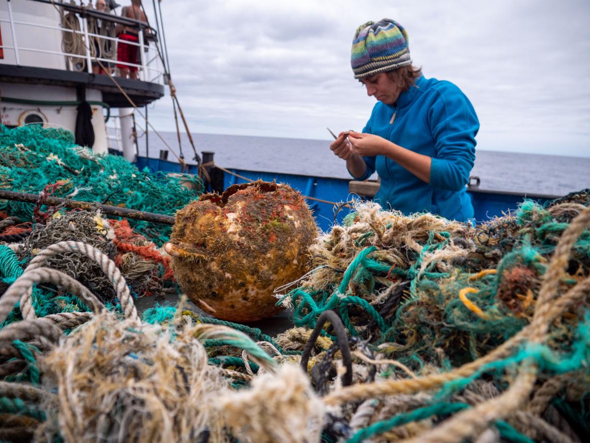 Scientist in a blue jacket and hat stands on a boat, inspecting a piece of debris from a large pile of nets, rope and plastic