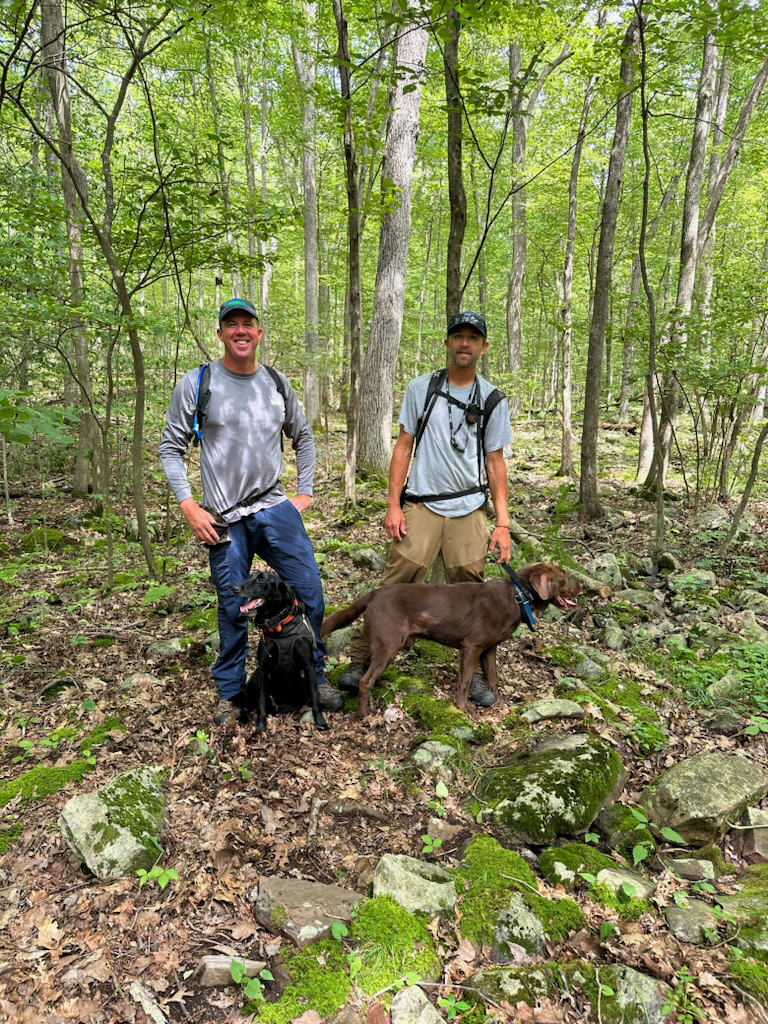 Two young men in caps stand in a forest, with a black Labrador and a chocolate Labrador in front of them.