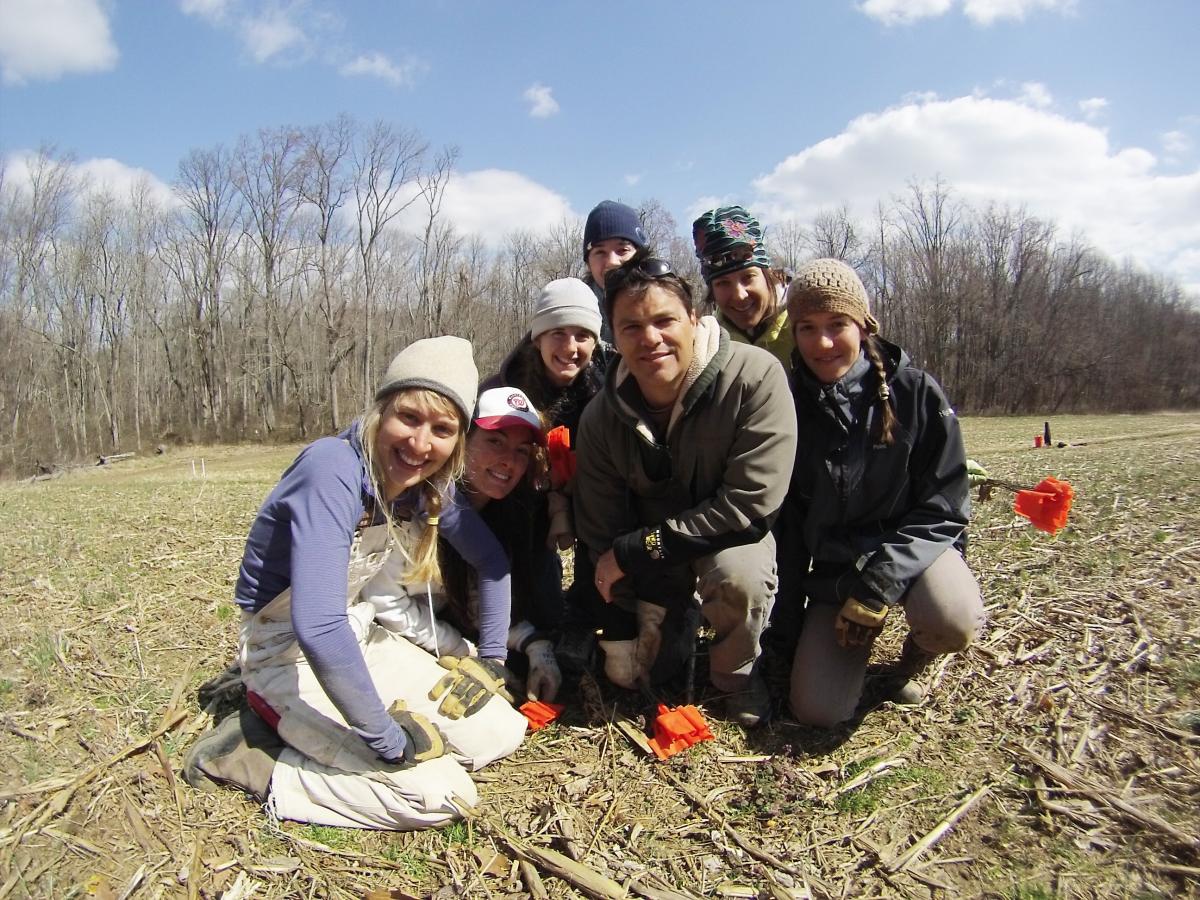 Seven young adults in winter clothes kneel for a photo on a grassy field