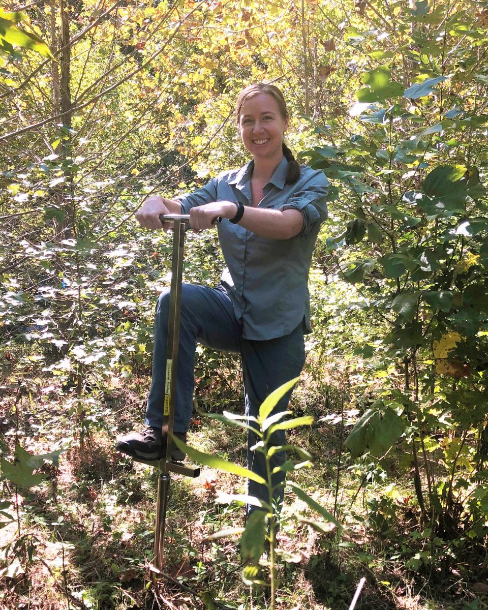 Young woman stands with one foot on a soil corer in a patch of trees