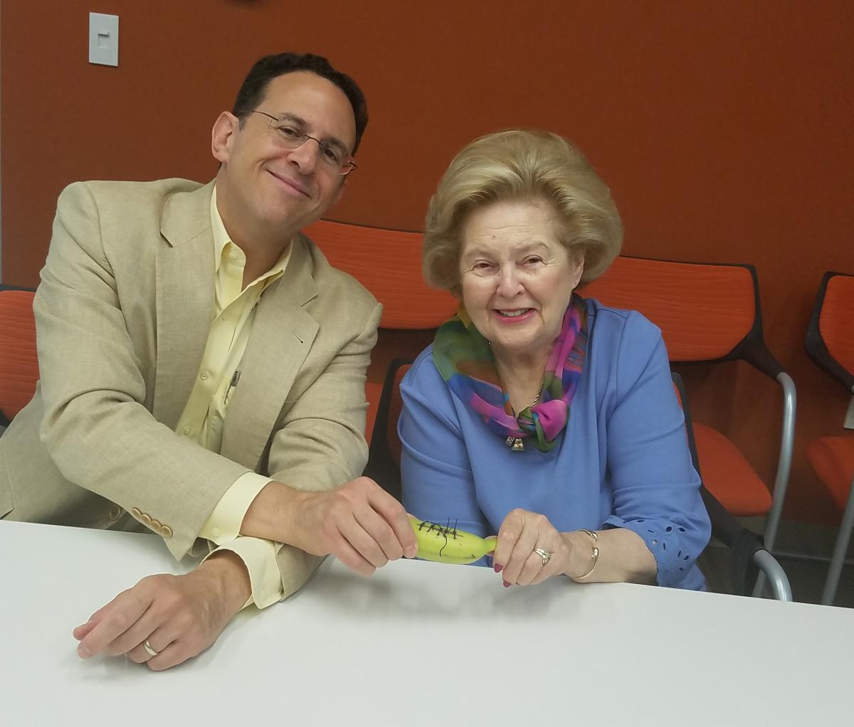 Two adults in suit jackets sit at a table, holding out a banana with black sutures on its side