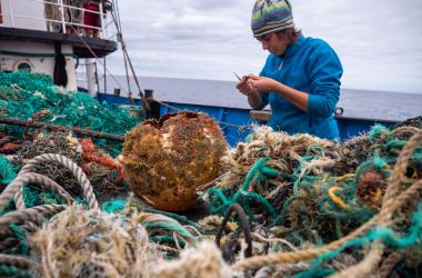 Scientist in a blue jacket and hat stands on a boat, inspecting a piece of debris from a large pile of nets, rope and plastic
