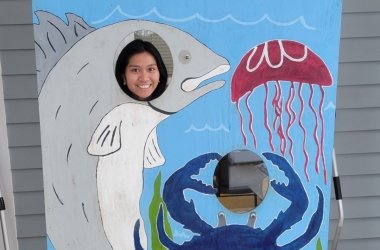 Julienne smiling while posing in a cut-outof a shark, crab, and jellyfish outside.