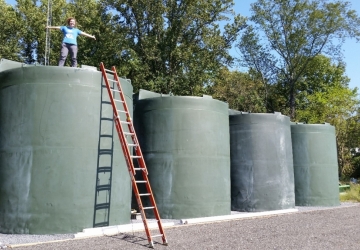 A woman stand on top of large treen tanks, against which is propped an extended orange ladder. 