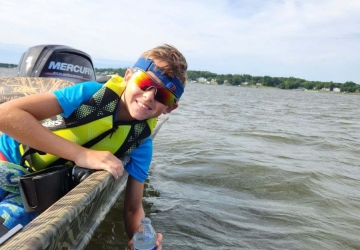 A young boy smiles facing the camera while he reaches over the edge of a small boat to get a water sample in a water bottle.