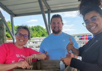 Three people smile at the camera and pose around a wooden stand for processing. They are on a dock on a sunny day.