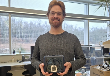 Volunteer smiles at the camera holding a camera in a lab with benches next to and behind him.