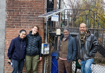 EJJI and SERC staff gather with community volunteer around the air quality sensor unit and smile at the camera.