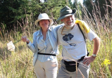 two people stand with their arms around each other on a sunny day in a field bordered by a forest. They smile at the camera.