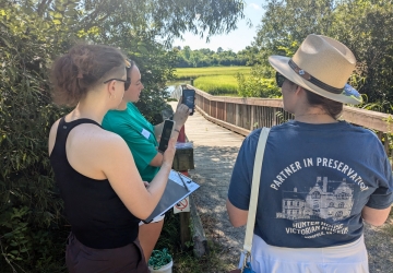 Rebecca holds up her phone to two people looking on. Backdrop is a sunny boardwalk leading to a marshy area.