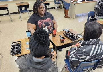 Project coordinator sits at desks with two students and is explaining to them how to set up the trays that are on the desks.