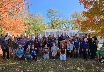 Volunteers gather as a group, standing and squatting, smiling. They are on grass in between two fall-color trees.