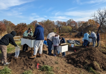 Volunteers sift through soil and stand around the screens and piles of sifted soil. They are against a forested backdrop and blu