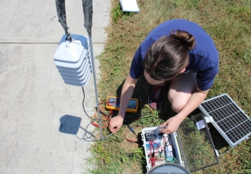 A white woman sets up the sensor's connections in a storage box that is resting on the grass.