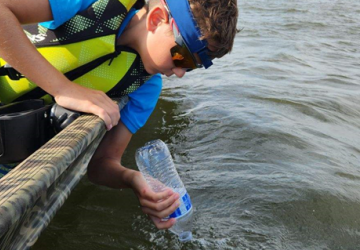 A boy in sunglasses and life vest on a boat gathers a water sample from dark blue-gray water using a water bottle.