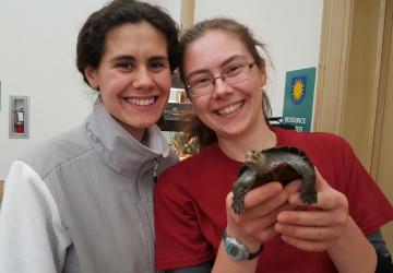 A young woman wearing a white & gray jacket poses next to a young woman in a red t-shirt & glasses holding a turtle