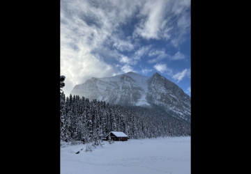 A small cabin in the snow sits on the edge of a forest, dwarfed by the giant gray mountain behind it.