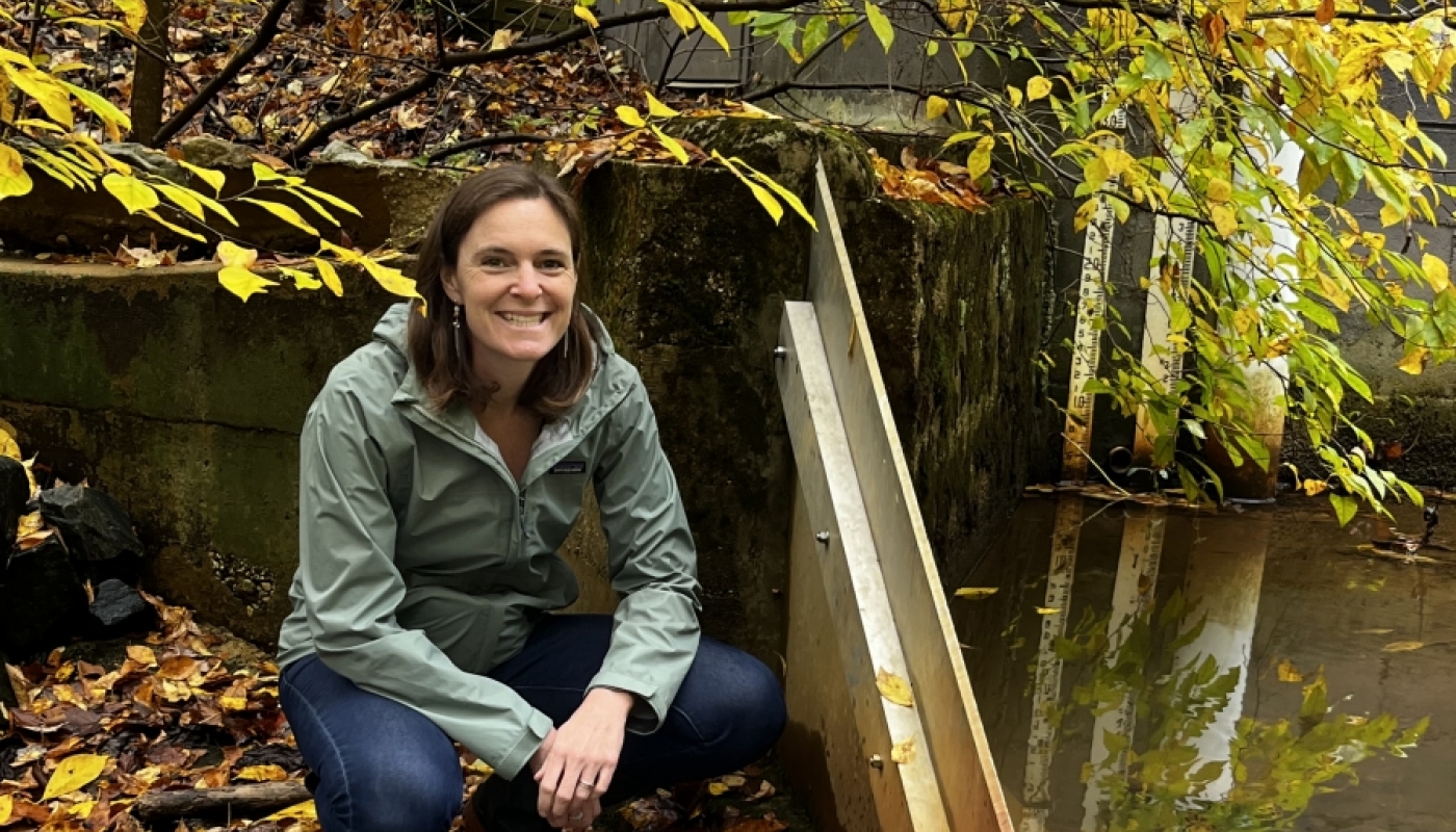 Rebecca Hale crouches in a stream while smiling at the camera, beside a V-shaped weir.
