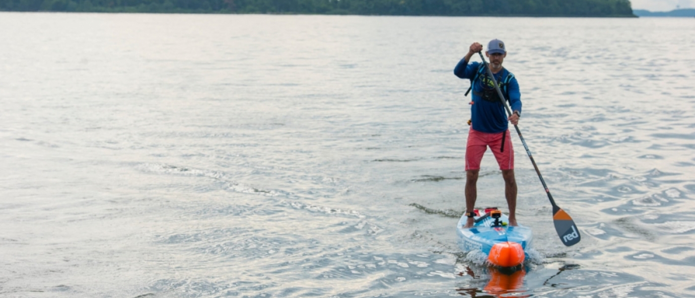 A man paddles a blue and orange stand-up paddle board across the water, with a forested shoreline in the background.